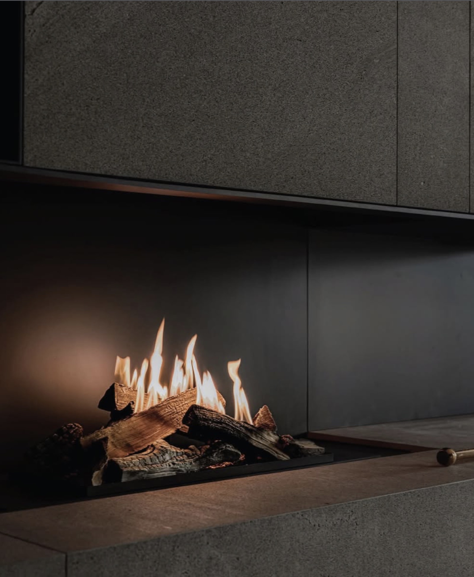 Close view of logs burning low in a linear fireplace set into a grey stone hearth.
